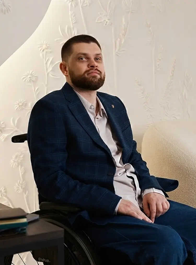 Мужчина в инвалидной коляске в синем костюме и голубой рубашке сидит в комнате с белыми стенами рядом со светлым диваном.