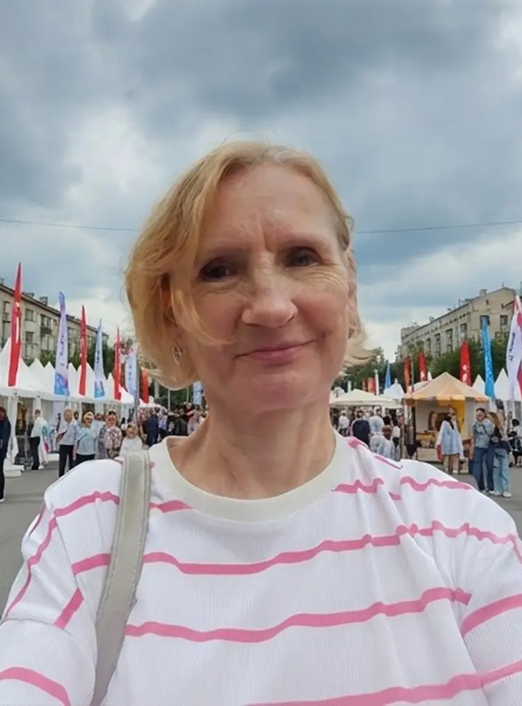 Женщина с короткими светлыми волосами в кофте в розовую полоску стоит на площади, рядом с ней много людей и красных флагов.