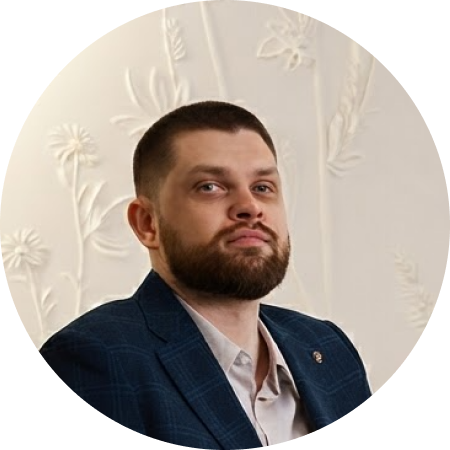 Мужчина в инвалидной коляске в синем костюме и голубой рубашке сидит в комнате с белыми стенами рядом со светлым диваном.