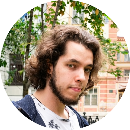 Мужчина с длинными темными волнистыми волосами и бородой в серой куртке и белой футболке с нарисованными серыми пальмами стоит напротив светлого здания, позади него находится черная резная ограда и деревья.