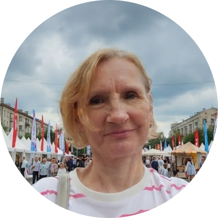 Женщина с короткими светлыми волосами в кофте в розовую полоску стоит на площади, рядом с ней много людей и красных флагов.